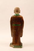 A CHINESE SANCAI-GLAZED MODEL OF A LOHAN. Ming Dynasty. Standing on a low rectangular base in long